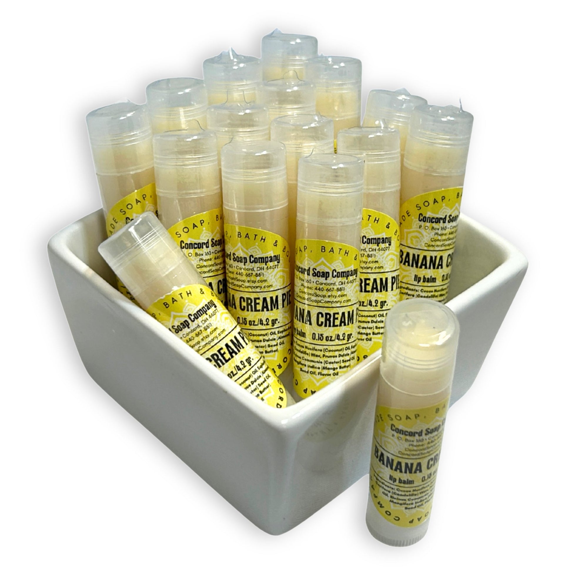 Banana Cream Pie handmade lip balm tubes in a white ceramic bowl, with one tube standing out front. This Banana Cream Pie natural lip balm by Concord Soap Company is labeled with a sunny yellow label that contains product and company details, and is then individually shrink wrapped for safety.