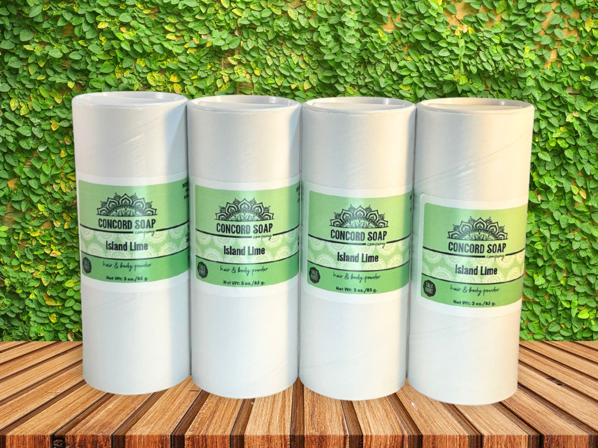 Four eco-friendly white cardboard shakers of handmade Island Lime hair and body powder by Concord Soap Company, displayed on a wood surface with a bright green leaf background. Natural talc-free powder in the coconut lime verbena fragrance for fresh, tropical scent skincare and personal hygiene.