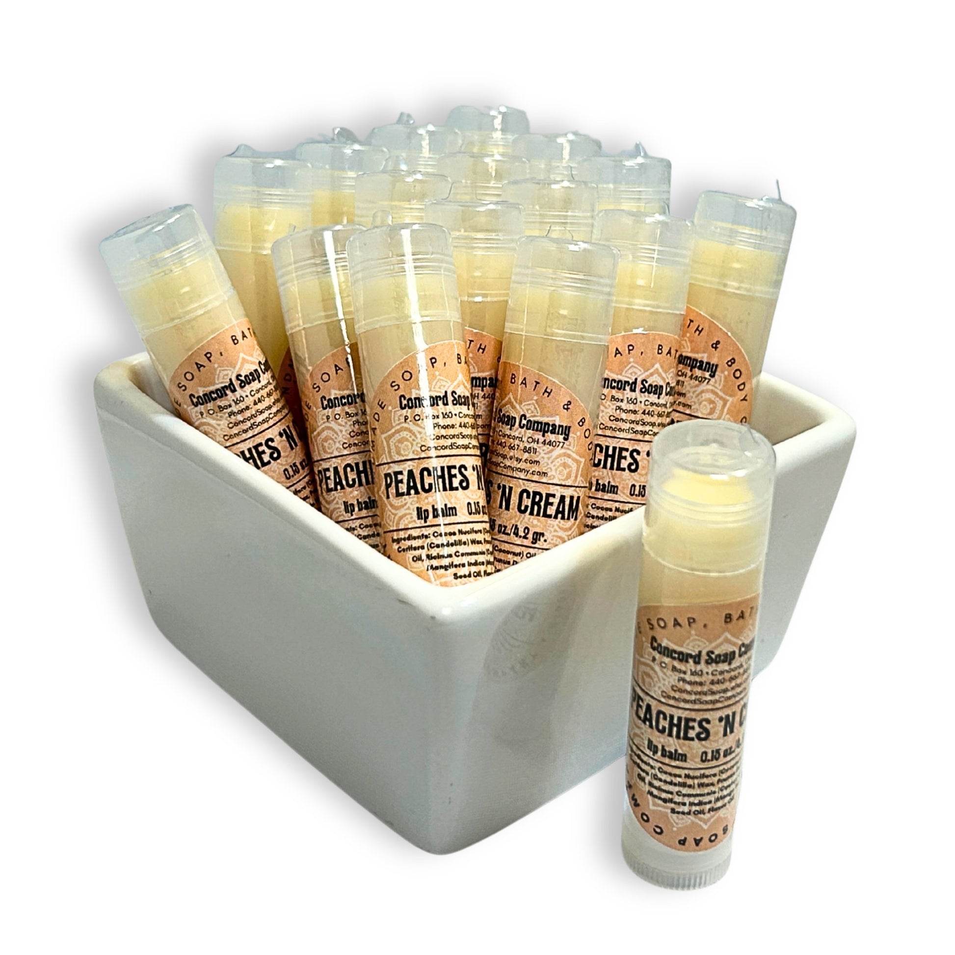 Peaches & Cream handmade lip balm tubes in a white ceramic bowl, with one tube standing out front. This Peaches & Cream natural lip balm by Concord Soap Company is labeled with a peach-colored label that contains product and company details, and is then individually shrink wrapped for safety.