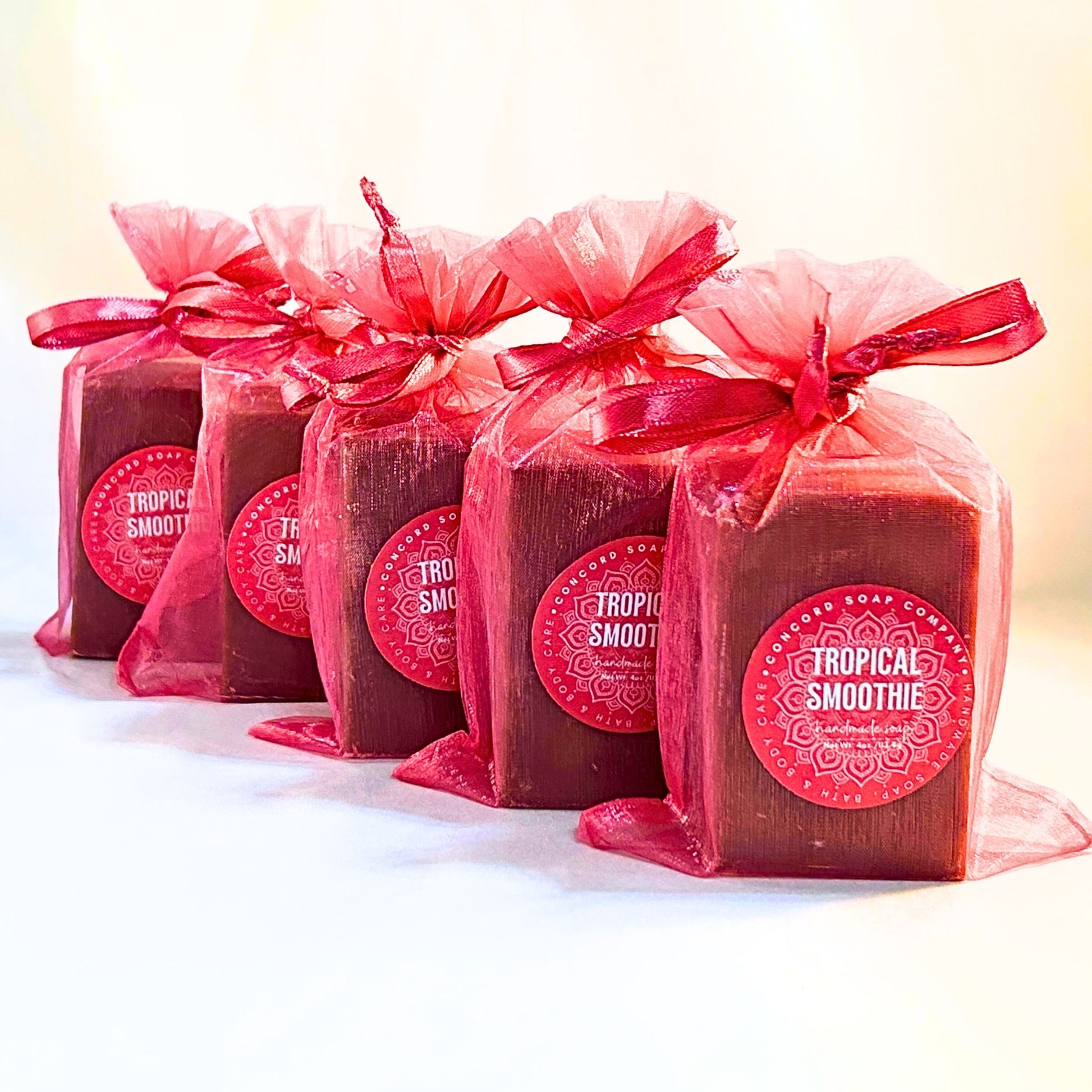 Five Tropical Smoothie handmade soap bars in bright pink organza gift bags with round Concord Soap Company labels, lined in a row. 