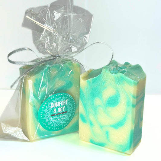 Clear cello wrapped bar of Comfort & Joy handmade soap with a teal and cream swirl design, tied with a silver ribbon, sitting next to an unwrapped bar.